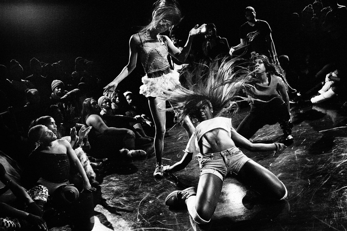 A black-and-white image shows a group of figures during a performance. The person in the foreground wears shorts and cropped shirt; they kneel on the ground with flailing hair while another person in a white skirt bends her head to one side. Other people stand around them and watch.